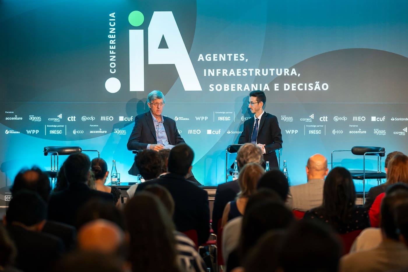Conferência .IA: dois oradores em palco, a plateia em contraluz.